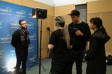 Young man standing in front of a blue background while he shares his views about what makes Fairfield unique and is filmed by three people