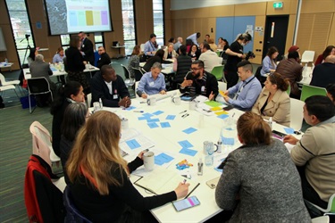 Group of people around a table listening and taking notes as one of the participants shares his views relating to their group activity