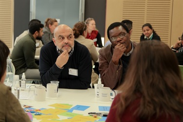 Photo of two men sitting at table touching their chins as they listen intently to someone speaking off camera at Fairfield Conversations summit