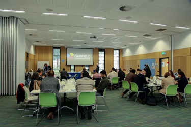 View of the room used for Fairfield Conversations summit, with summit attendees seated around a number of tables and a projected image on the wall with the Fairfield Conversations logo