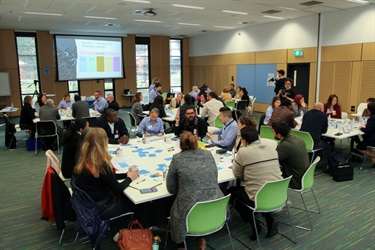 four tables of people working on group activities at the Fairfield Conversations summit, while a PowerPoint slide is projected onto a projector screen at the end of the room