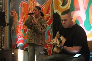 Local artist, Jessica Jade, singing into a microphone, next to a guitarist, at Bonnyrigg Youth Centre. Photographed by Hannah Teau & Noor Abu-Sada