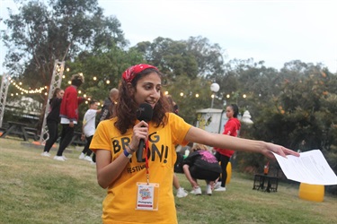 Young volunteer speaking into a microphone and gesturing to the side while holding a piece of paper, in the outdoor area of the Bonnyrigg Youth Centre. Photographed by Hannah Teau & Noor Abu-Sada