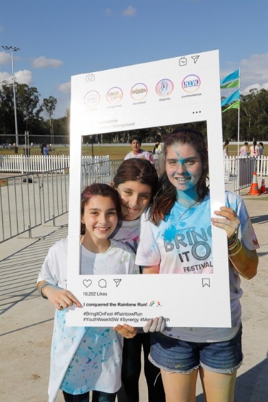 Three young friends covered in colour powder and posing with Instagram frame prop