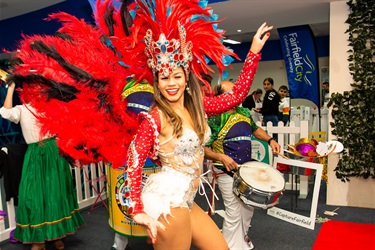 Brazilian dancer, wearing red and white costume, smiling and dancing in front of man playing the drums