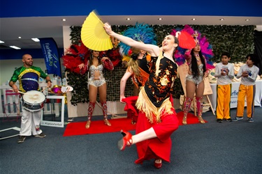 Young women wearing traditional Spanish dresses and holding colourful fans, performing alongside a drummer and Brazilian dancers
