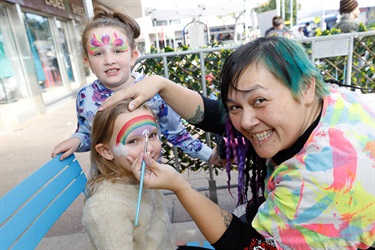 Face painter smiling and posing while painting rainbow face paint on young girl