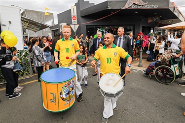 Two young men wearing sport jerseys carrying and hitting drums while leading parade of young children