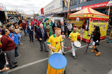 Two young men in sport jerseys and hitting drums leading parade of Councillors, young children and Brazilian dancers