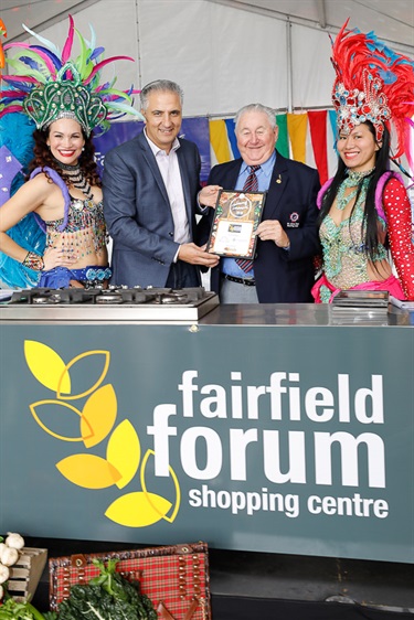 Mayor Frank Carbone and Saint John's Bowling Club representative holding a certificate while posing with two female Brazilian dancers