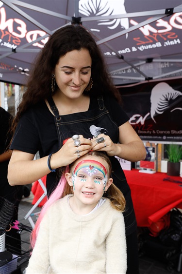 Young woman smiling while braiding hair of young girl with rainbow face paint