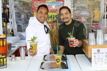 Two men smiling and posing while holding yellow and pink sugar cane juice drinks