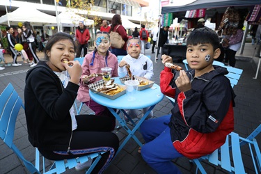 Four young children with colourful face paint smiling and posing while sitting around a table and eating meat skewers