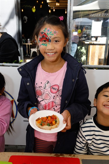 Young girl with lady bug face paint smiling and posing while holding a plate of bread with sliced cherry tomatoes
