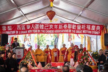Man at a podium speaking into a microphone while a group of monks sit on stage behind a table with plates of traditional Chinese food