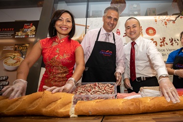 Mayor Frank Carbone smiling and posing with Councillor Dai Le and Adrian Wong while preparing the Vietnamese bread roll
