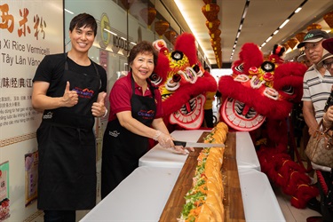 Andy Trieu and elderly baker smiling and posing with red lion dancing puppets while holding a bread knife to the Vietnamese bread roll