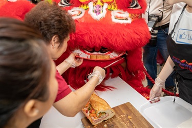 Elderly baker placing portion of Vietnamese bread roll into the mouth of a red lion dancing puppet