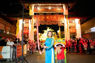 Dai Le in traditional Vietnamese Ao dai lighting fire crackers in front of a large crowd at Pai Lau gate with a child wearing a lion dancers lion head next to her