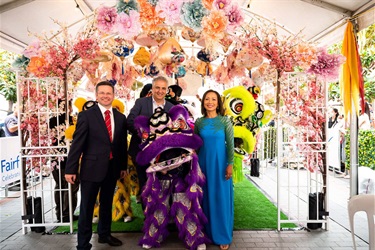 Mark Coure, Frank Carbone and Dai Le smiling and posing with a child lion dance performer in a purple lion costume underneath a colourful lantern garden decoration with flowers and lights