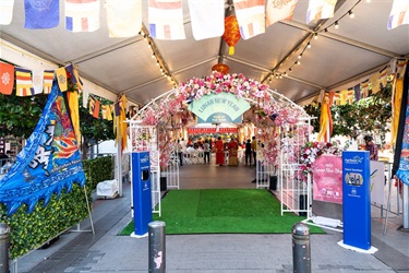 A white frame decorated with assorted pink, red, white and yellow flowers and colourful lanterns. A green Lunar New Year fan sign hangs on the front of the frame. The frame is underneath a white marquee decorated with Buddhist flags and lights.