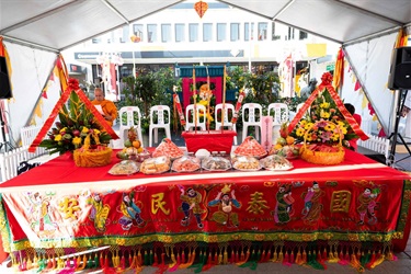 A long table with a red cloth. The cloth has Chinese lettering, colourful art of people dancing and playing traditional musical instruments. On the table are plates of food, flower decorations and a large incense holder with incense that are lit.