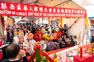 A large crowd surrounding and underneath a decorated marquee. At the front of the crowd are colourful lion dance performers.