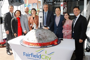 New South Wales Premier Gladys Berejiklian and Mayor Frank Carbone speaking into a microphone while standing next to table holding a cake decorated to be astronaut and rocket standing on the moon