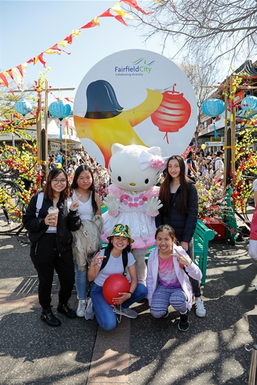 Group of young friends smiling and posing with Hello Kitty costumed character in front of the Cabramatta Moon Festival banner
