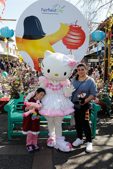 Woman and young girl smiling and posing with Hello Kitty costumed character in front of Cabramatta Moon Festival banner