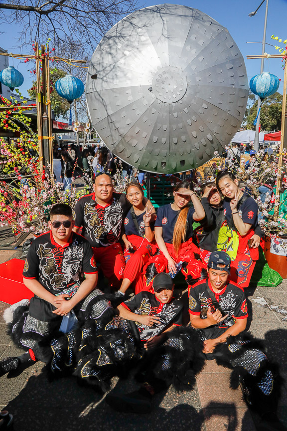 Team of lion dancers wearing red and black uniform smiling and posing in front of Cabramatta Moon Festival decorations