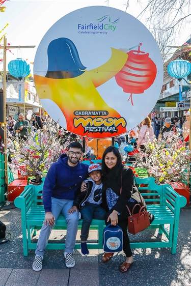 Family smiling and posing while sitting on a teal bench under Cabramatta Moon Festival banner