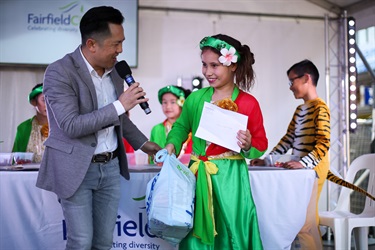 Man speaking into a microphone presenting bag and envelope to young girl wearing traditional cultural dress