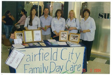 Fairfield City Family Day Care educators, wearing uniform, smiling and posing at their stall holder table at Wetherill Park Stocklands