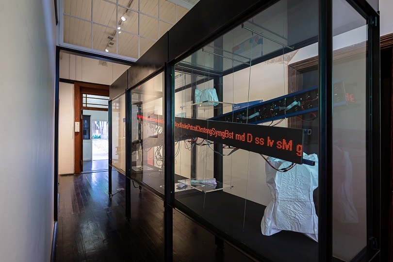 Interior of a museum with a glass display case. In the case there is red LED text visible and objects wrapped in white material. 