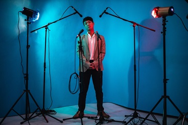Studio portrait of a man standing in the spotlight against a bright blue wall with microphones and lights pointing at him