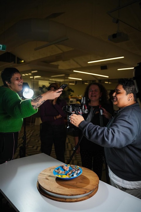 A group of people are gathered around a table with a wooden turntable and a plate of food on it. One person is holding a light, another is holding a microphone, and another is operating a camera.