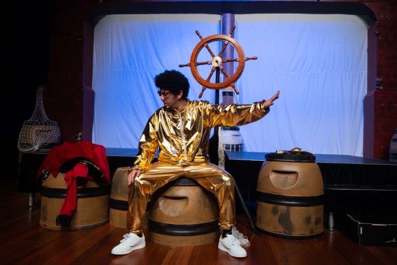 A person dressed in a shiny gold outfit and white sneakers is sitting on a barrel with one arm extended to the side. The background features a large ship's wheel mounted on a blue and white backdrop.
