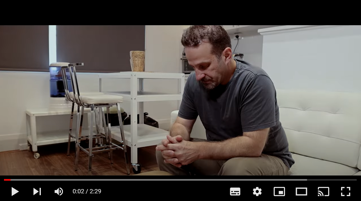 Screenshot of opening scene of a YouTube video with a man sitting on a white lounge with his head down and arms resting on his legs with hands closed together