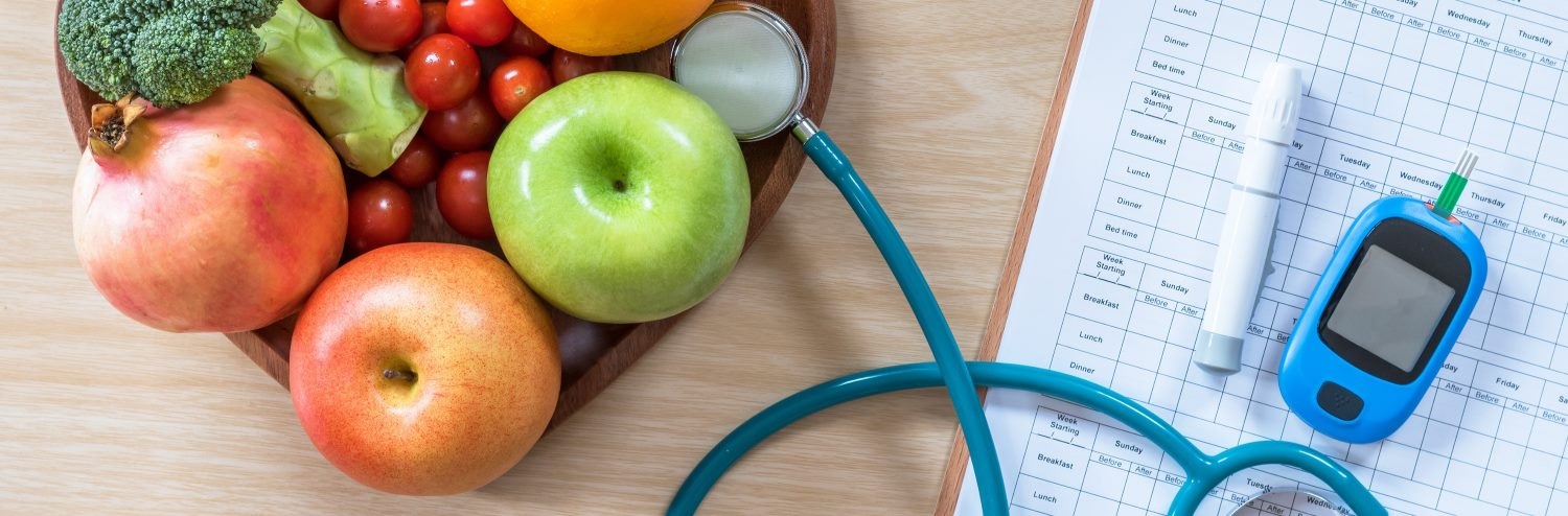 A wooden table displaying a wooden bowl filled with apples, oranges, tomatoes, and broccoli. Beside the bowl are a stethoscope, a blood glucose meter with a test strip, an insulin pen, and a clipboard with a medical chart.