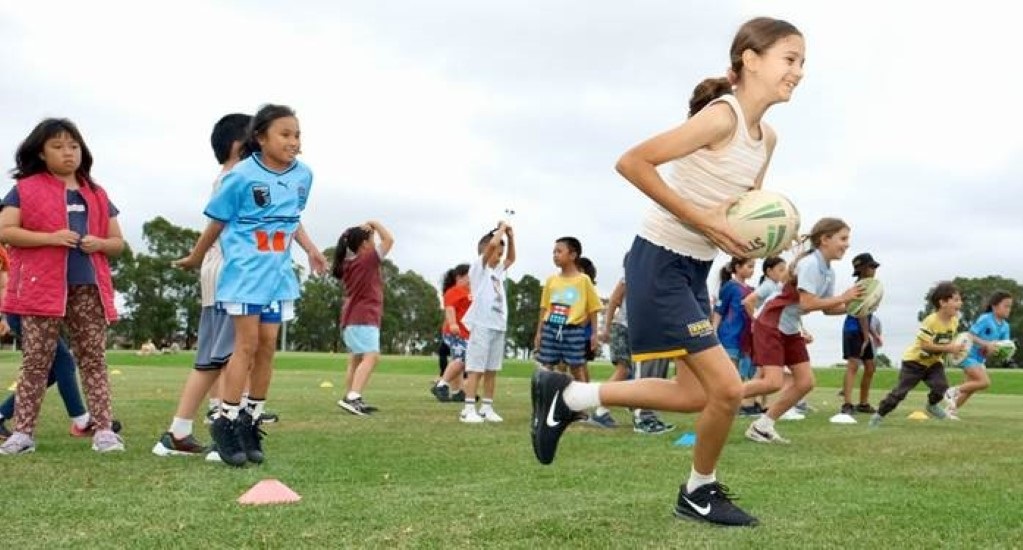 A group of young girls joyfully playing football on a grassy field under a clear blue sky.