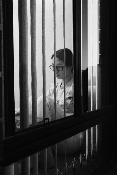 Black and white photo of person at home using a lap top computer, viewed through her window and blinds from outside her house at night time