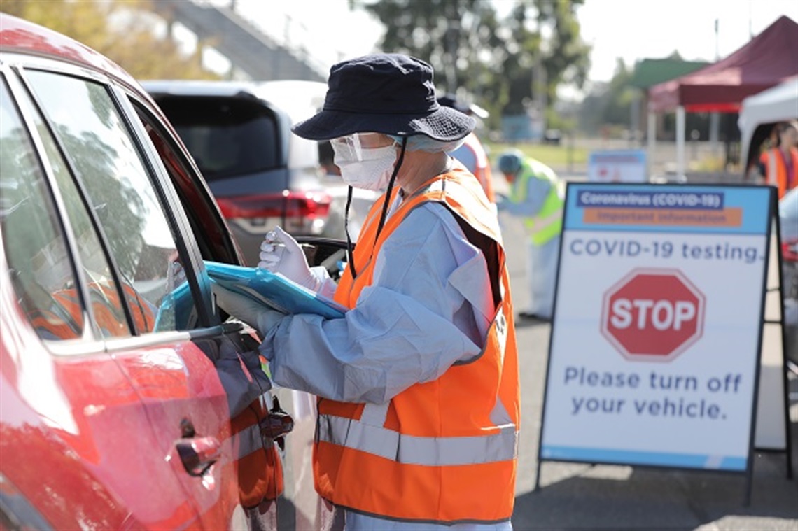 Lady in mask and gloves at red car with clipboard with COVID testing sign in background 