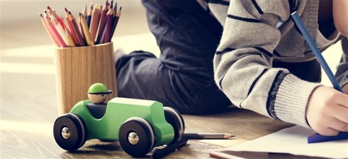 A young boy drawing with coloured pencils next to a wooden green toy car