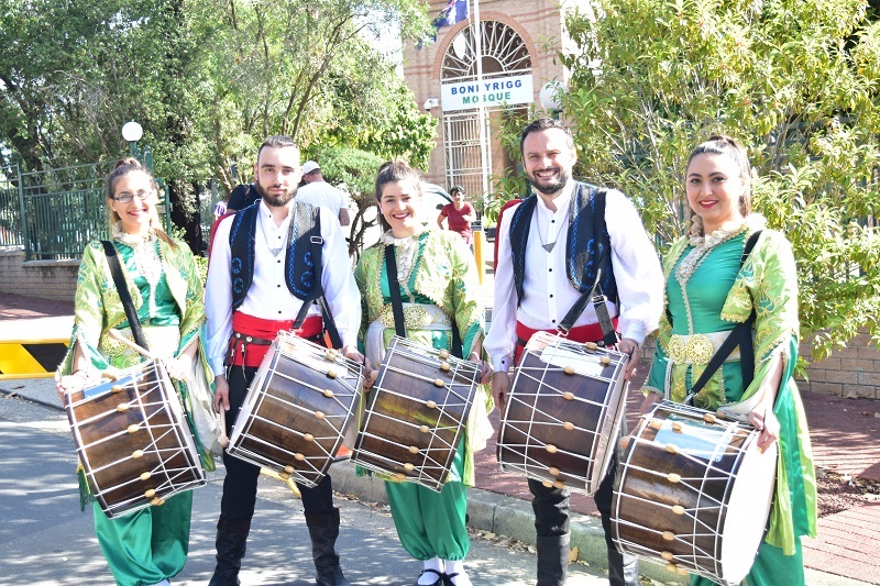Members of the Bonnyrigg Mosque dance group, wearing traditional cultural attire and holding drums, smiling and posing in front of the Bonnyrigg Mosque