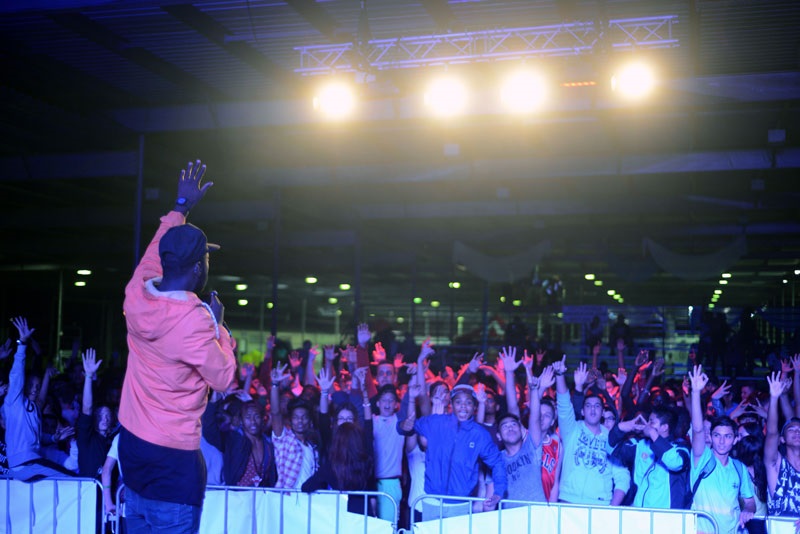 Performer speaking into a microphone, to a crowd of young people dancing with their arms in the air, at the Bring It On! Festival