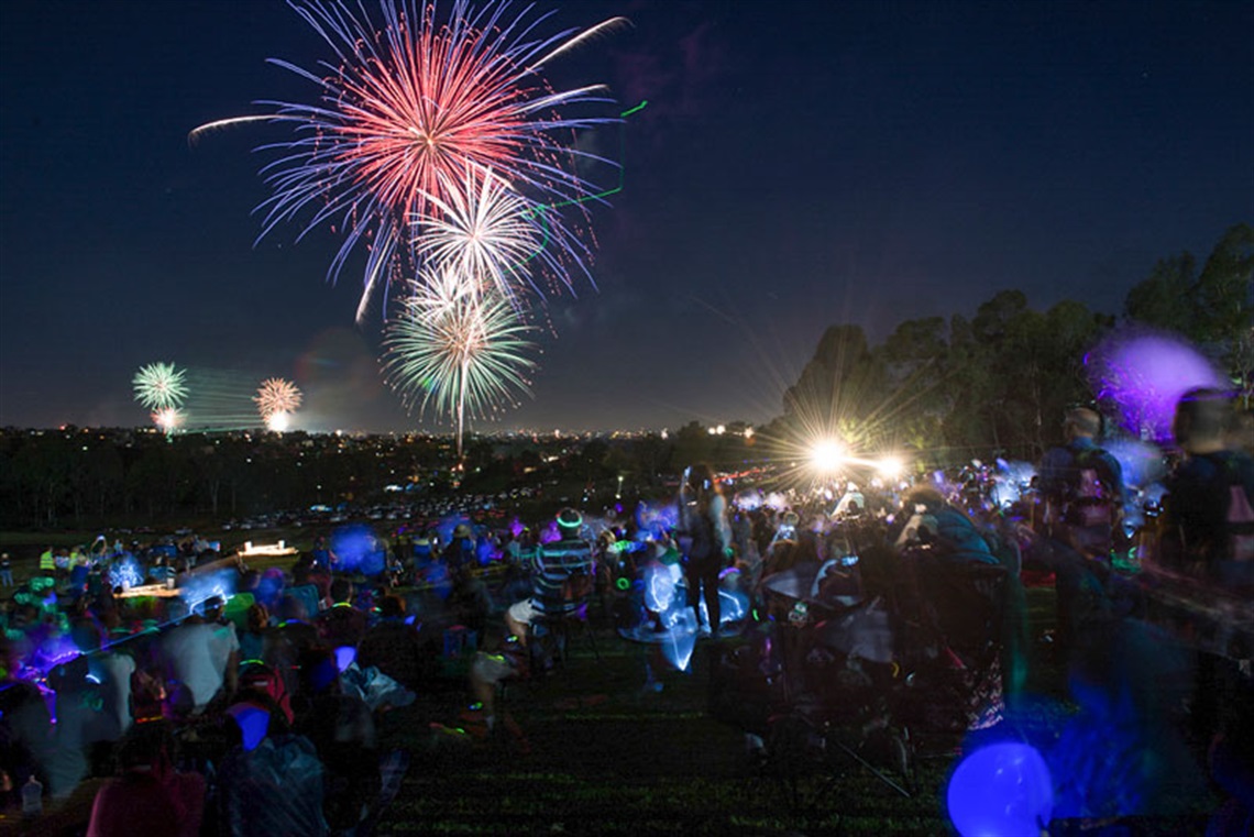 Crowd of guests seated on a field, watching the spectacular display of green, red, blue, orange and white fireworks 