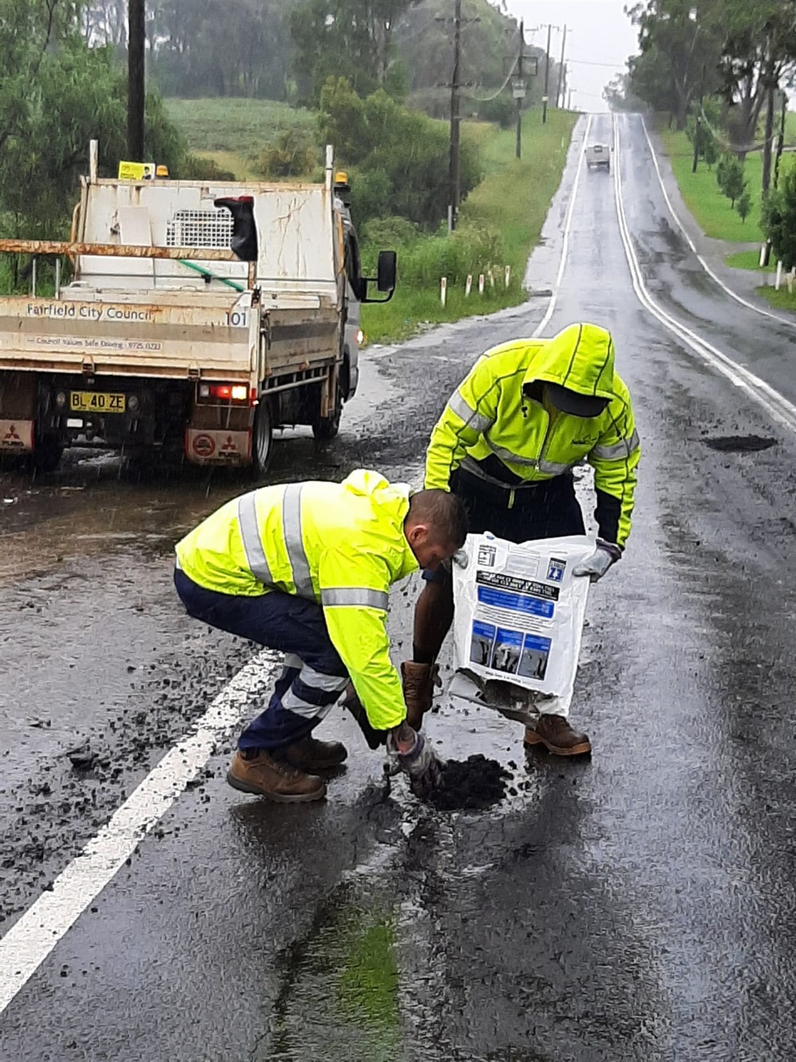 Two men wearing high visibility jackets filling in pot hole on the road  