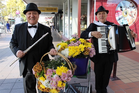 Two men wearing suits playing the clarinet and accordion while holding baskets of yellow, pink and white flowers