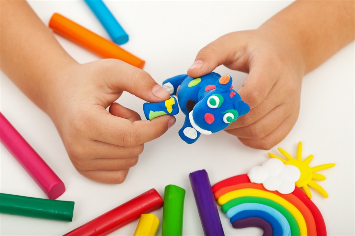 A child's hands forming an animal sculpture out of blue clay above colourful pieces of clay and rainbow sculpture